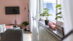 A living room with a TV mounted on the wall, a window with a view, a radiator, and a potted plant on the windowsill.