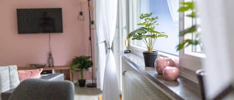 Un salon avec un téléviseur fixé au mur, une fenêtre offrant une vue agréable, un radiateur et une plante en pot posée sur le rebord de la fenêtre.