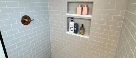 A shower with a glass door, a white tiled floor, and a shelf with toiletries.