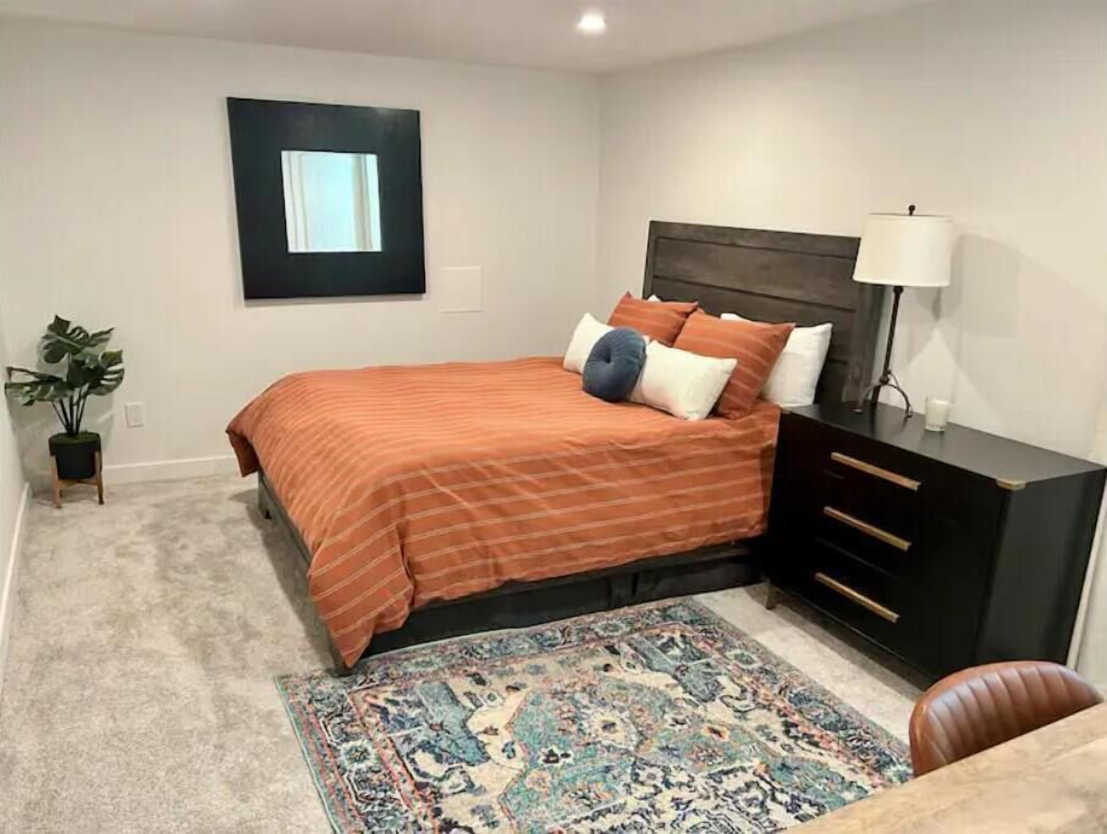 Un dormitorio con cama, mesita de noche, lámpara, planta en maceta y alfombra.