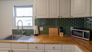 Une cuisine avec des meubles blancs, un plan de travail en bois, un évier en acier inoxydable, un four à micro-ondes et une fenêtre donnant sur un bâtiment à l’extérieur.