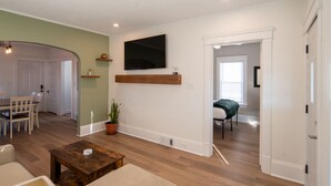 Un vaste salon doté de planchers en bois franc, d’un téléviseur à écran plat fixé au mur, d’un coin repas avec une table et des chaises, et d’un espace salon confortable avec une table basse.