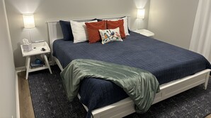 A neatly made bed with a blue quilt, white headboard, and multiple pillows. There are two bedside tables with lamps, a small shelf with books, and a black textured rug on the floor.