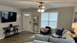 A living room with a grey sofa, a black armchair, a TV, a ceiling fan, and a window with blinds.