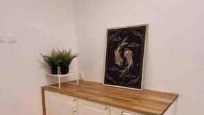 Een kleine, overzichtelijke kamer met een houten vloer, een witte kast met lades, een klein plankje met planten en een ingelijst kunstwerk aan de muur.