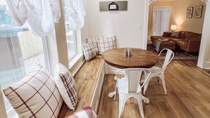 A cozy living room with a wooden floor, a round dining table, white chairs, a brown sofa, and a bench with plaid cushions.