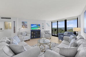 A modern living room with a large sofa set, a coffee table, and a television. There is a balcony with a view of the landscape.