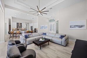 A modern living room with a sectional sofa, a coffee table, and a dining area with chairs and a kitchen in the background. - 2024 Monsterrat Model | Waterfront Home & Lap Pool (Marco Island)