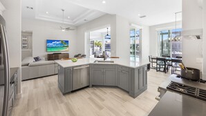 A modern kitchen with grey cabinets, a large island, and a dining area with a view. - 2024 Monsterrat Model | Waterfront Home & Lap Pool (Marco Island)