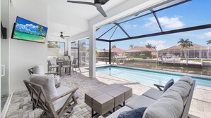 A modern indoor pool area with a view of a marina, including lounge chairs, a table, and a ceiling fan. - 2024 Monsterrat Model | Waterfront Home & Lap Pool (Marco Island)
