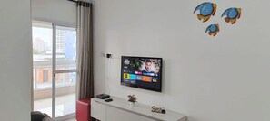 A room with a television mounted on the wall, a small white console table with a vase, and a window with curtains. - Residencial Octávio Braga (Praia Grande)