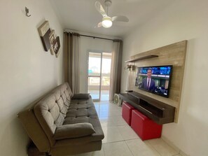 A living room with a sofa, a television mounted on a wooden wall, a ceiling fan, and a sliding glass door. - Residencial Octávio Braga (Praia Grande)