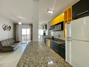 A modern kitchen with a central island, a dining area with a sofa, and a balcony with curtains. - Residencial Octávio Braga (Praia Grande)
