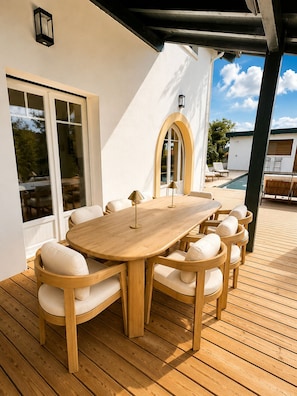 A wooden deck with a round table and chairs, a covered area with a pergola, and a pool in the background. - Magnifique Maison Moderne à Bidart - 15min à Pieds du Centre et de la Plage (Bidart)