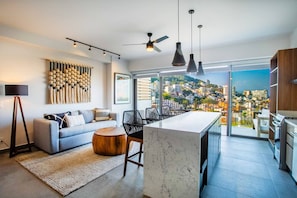A modern living room with a sofa, coffee table, and a dining area with chairs. There is a large window offering a city view. - Two Pools, Bar & Gym | Casa Medianoche at Oasis (Puerto Vallarta)