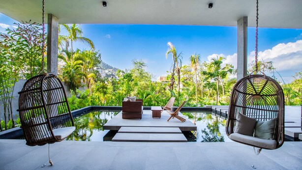 A serene outdoor seating area with two hanging chairs, a small table, and a bench, overlooking a lush garden and a body of water. - Two Pools, Bar & Gym | Casa Medianoche at Oasis (Puerto Vallarta)