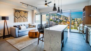A modern living room with a sofa, coffee table, and a city view through large windows. - Two Pools, Bar & Gym | Casa Medianoche at Oasis (Puerto Vallarta)
