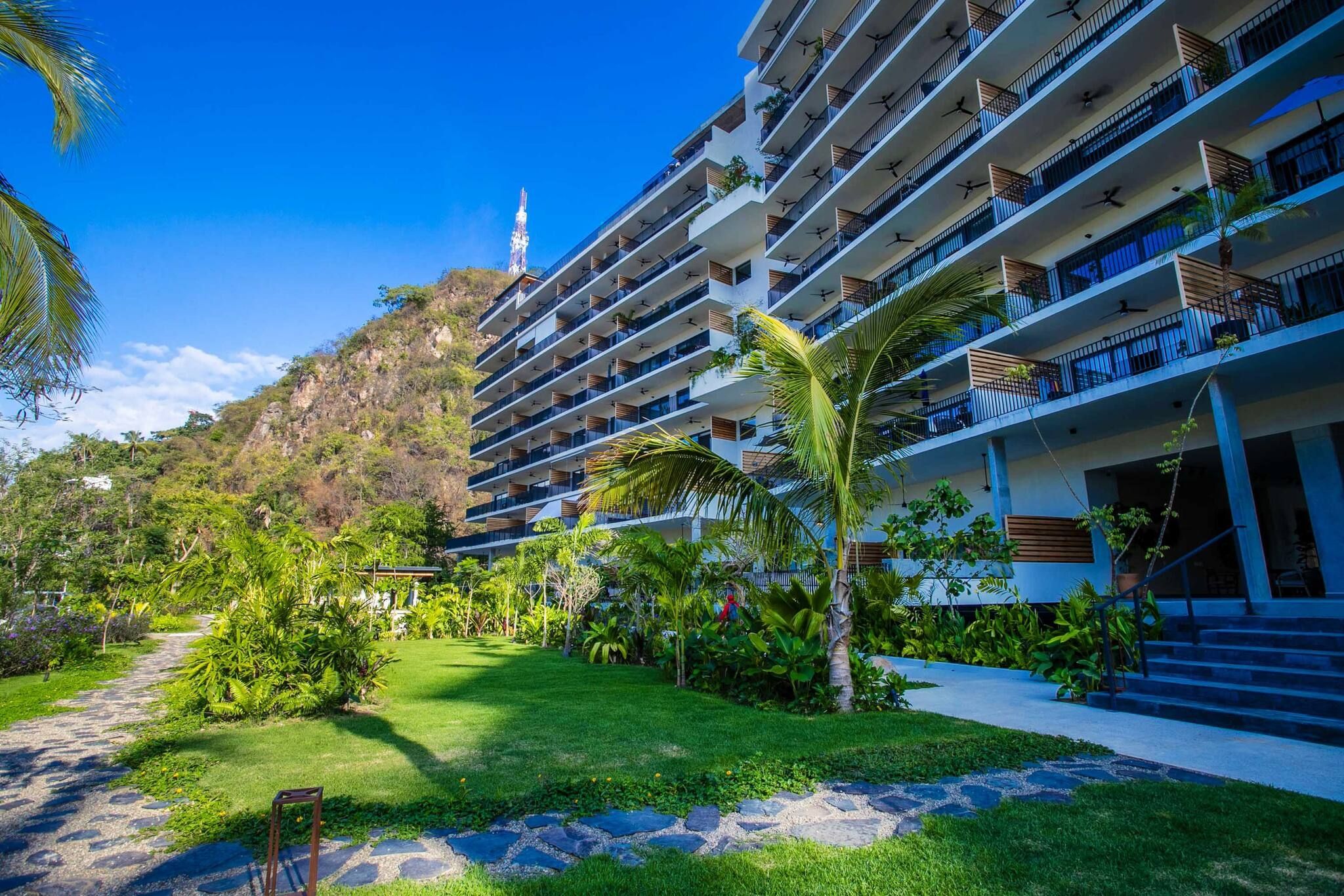 Un hotel de varios pisos con balcones, un jardín con plantas tropicales y un cielo despejado.