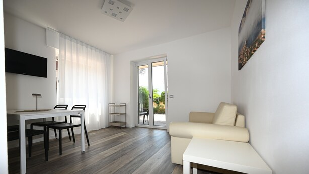 A modern living room with a sofa, a dining table with chairs, a TV mounted on the wall, and a sliding glass door leading to an outdoor area.