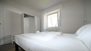 A neatly made bed in a room with a window, a radiator, and a coat rack.