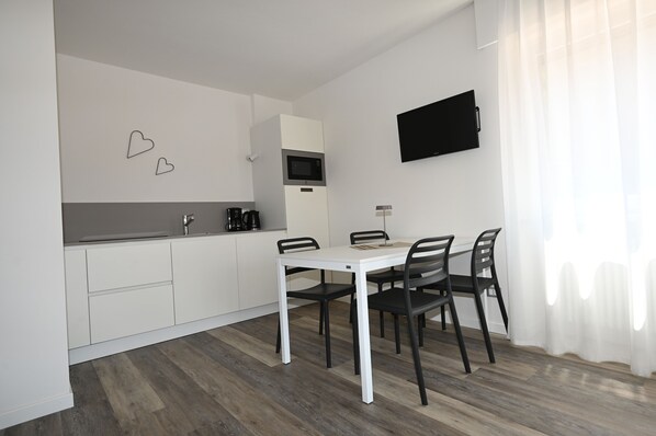 A modern kitchen with white cabinets, a white dining table with four chairs, a flat-screen TV mounted on the wall, and a coffee maker on the counter. - MODERN APARTMENT GROUND FLOOR AND PARKING SPACE APP WATER (Malcesine)