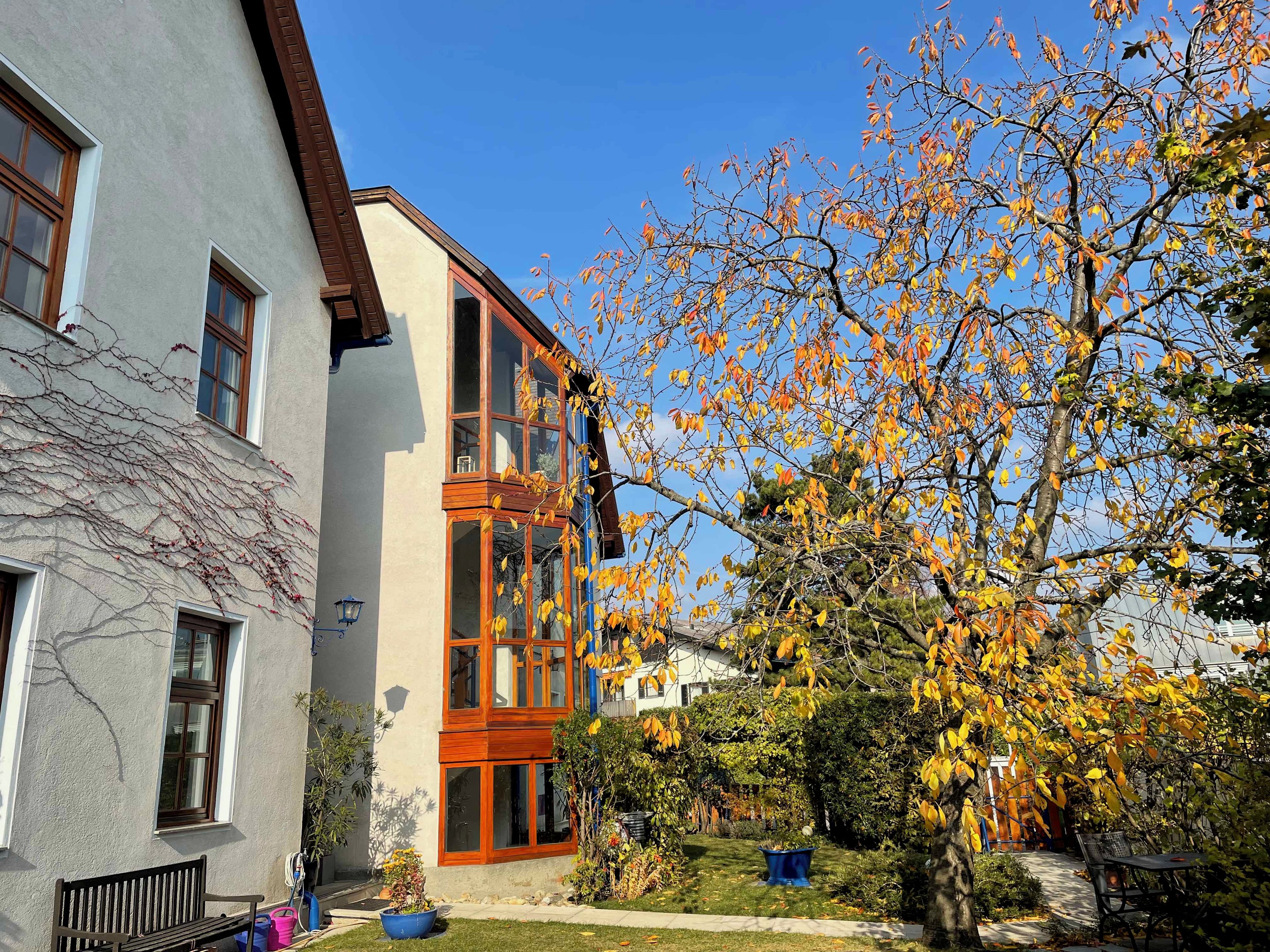 A two-story building with a balcony, a garden with a bench, and a tree with autumn leaves.
