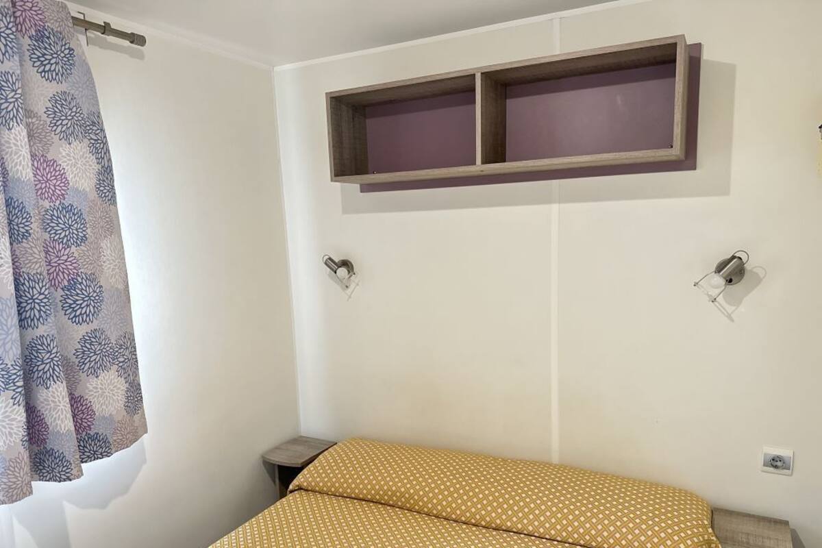 A bedroom with a bed, a window with a patterned curtain, a wall-mounted shelf, and a bedside table.