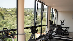 Un gimnasio con bicicletas estáticas y cintas de correr, amplias ventanas con vistas a zonas verdes y edificios, y suelo de madera.