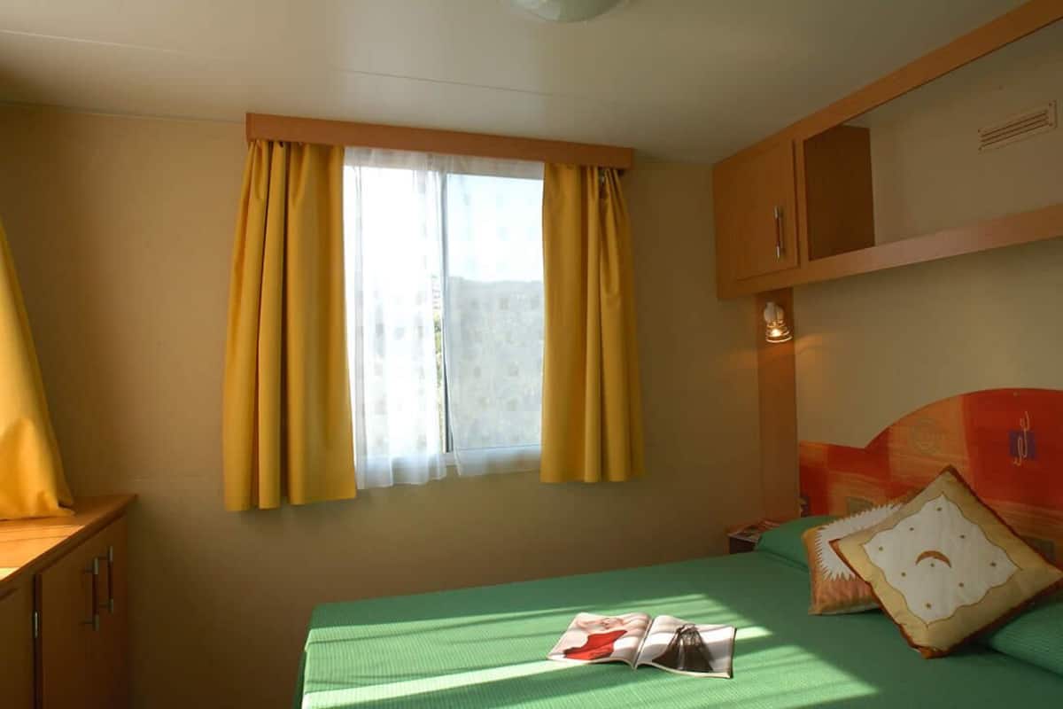 A room with a bed, a window with yellow curtains, a wooden cabinet, and a green bedspread.
