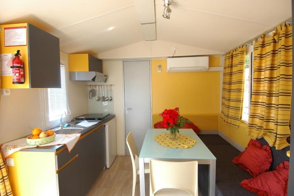 A compact kitchen with a sink, stove, and a small refrigerator. There is a dining area with a table and chairs, and a sofa with red cushions. A fire extinguisher is mounted on the wall above the kitchen.