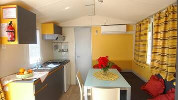A compact kitchen with a sink, stove, and a small refrigerator. There is a dining area with a table and chairs, and a sofa with red cushions. A fire extinguisher is mounted on the wall above the kitchen.