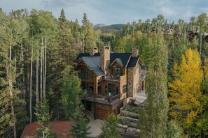 A large, multi-story log cabin with a prominent balcony, surrounded by dense forest and mountains in the background. - Modern Cabin by InvitedHome | Vast Patio, Fireplaces, Steps to Ski (Mountain Village)