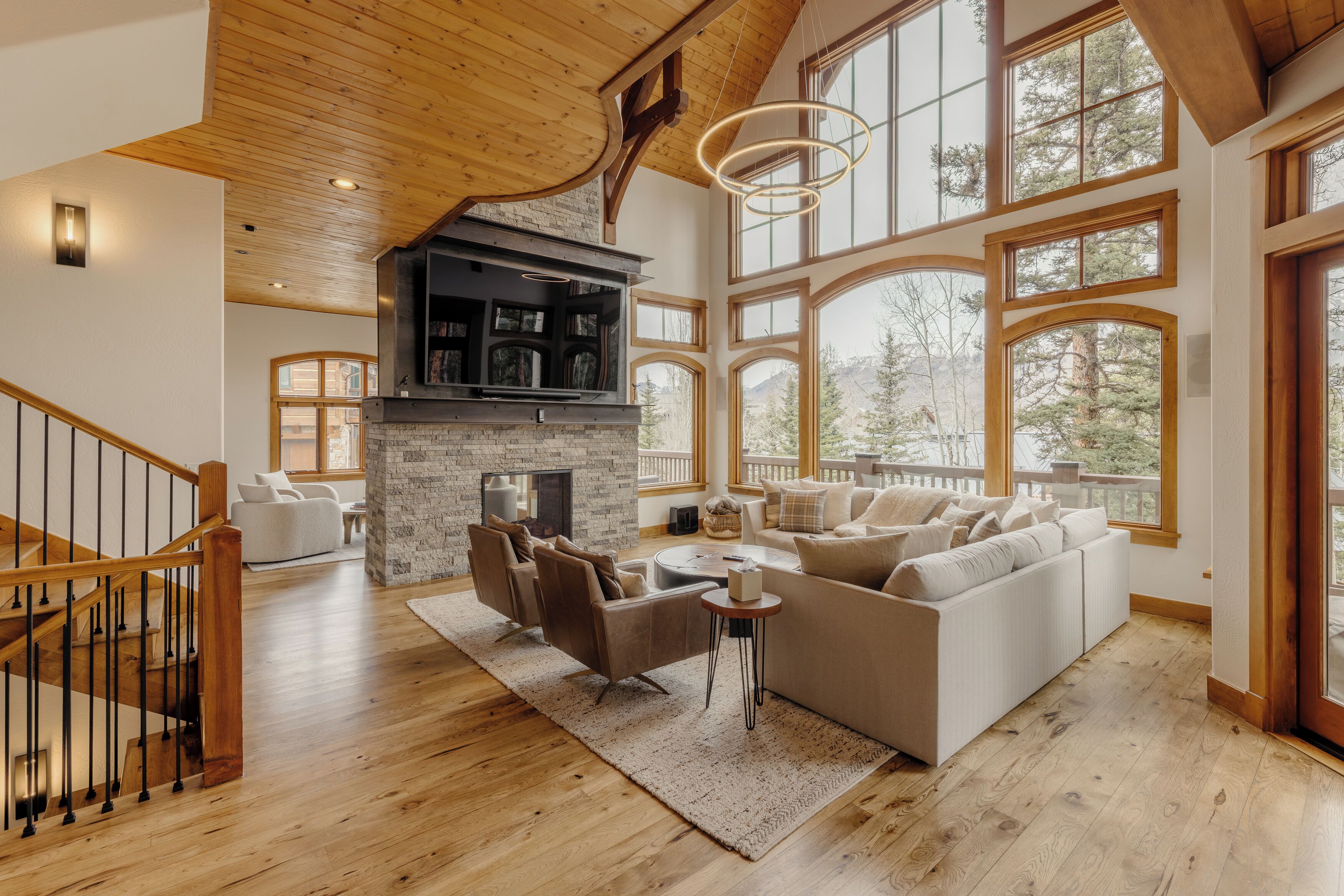 A spacious living room with a fireplace, large windows, and a wooden staircase.