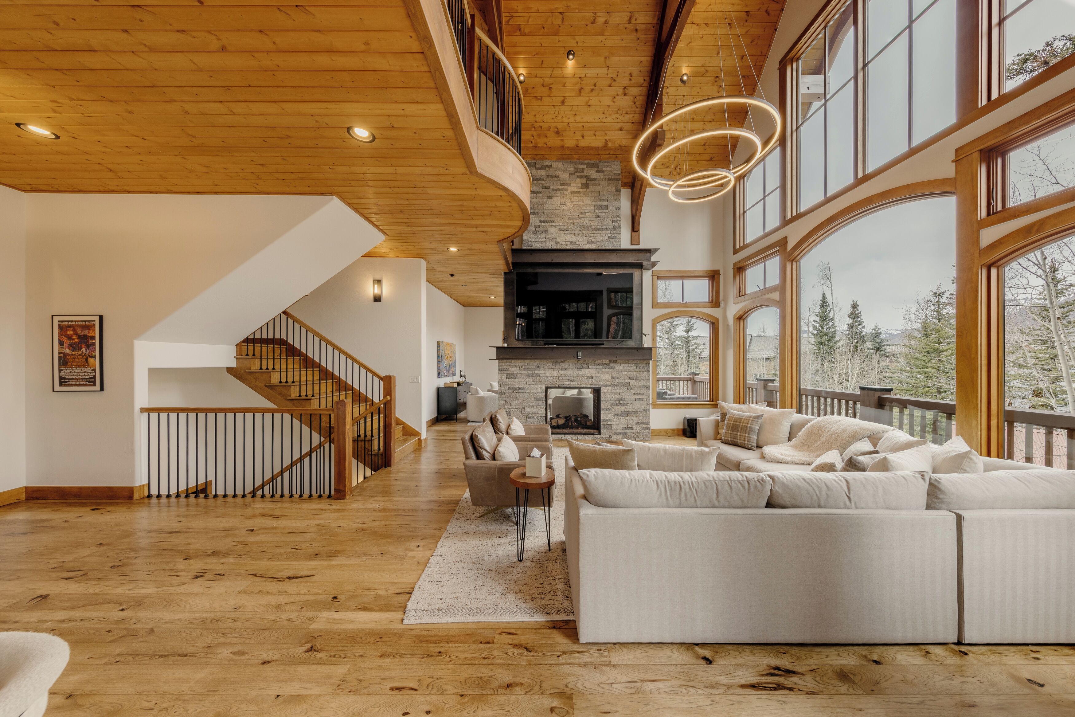 A spacious living room with a fireplace, large windows, and a wooden staircase.