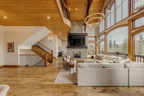 A spacious living room with a fireplace, large windows, and a wooden staircase. - Modern Cabin by InvitedHome | Vast Patio, Fireplaces, Steps to Ski (Mountain Village)
