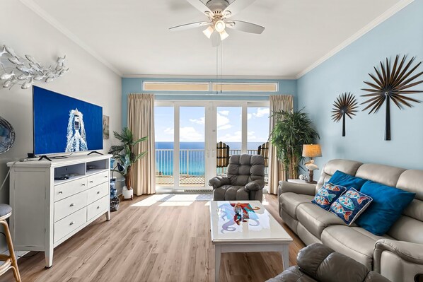 A living room with a sofa, coffee table, and a TV on a white cabinet. There is a balcony with a view of the ocean.