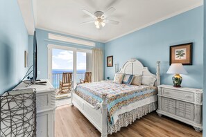 A bedroom with a bed, a nightstand, a desk, a chair, and a balcony with a view of the ocean.