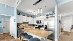 A modern kitchen with white cabinets, a granite island with bar stools, stainless steel appliances, and decorative wall art. - Gulf views w/ resort pools & beach access (Panama City Beach)