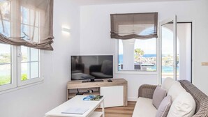 Una sala de estar moderna con un televisor de pantalla plana, una mesa de centro blanca, un sofá de mimbre y ventanas amplias con cortinas.