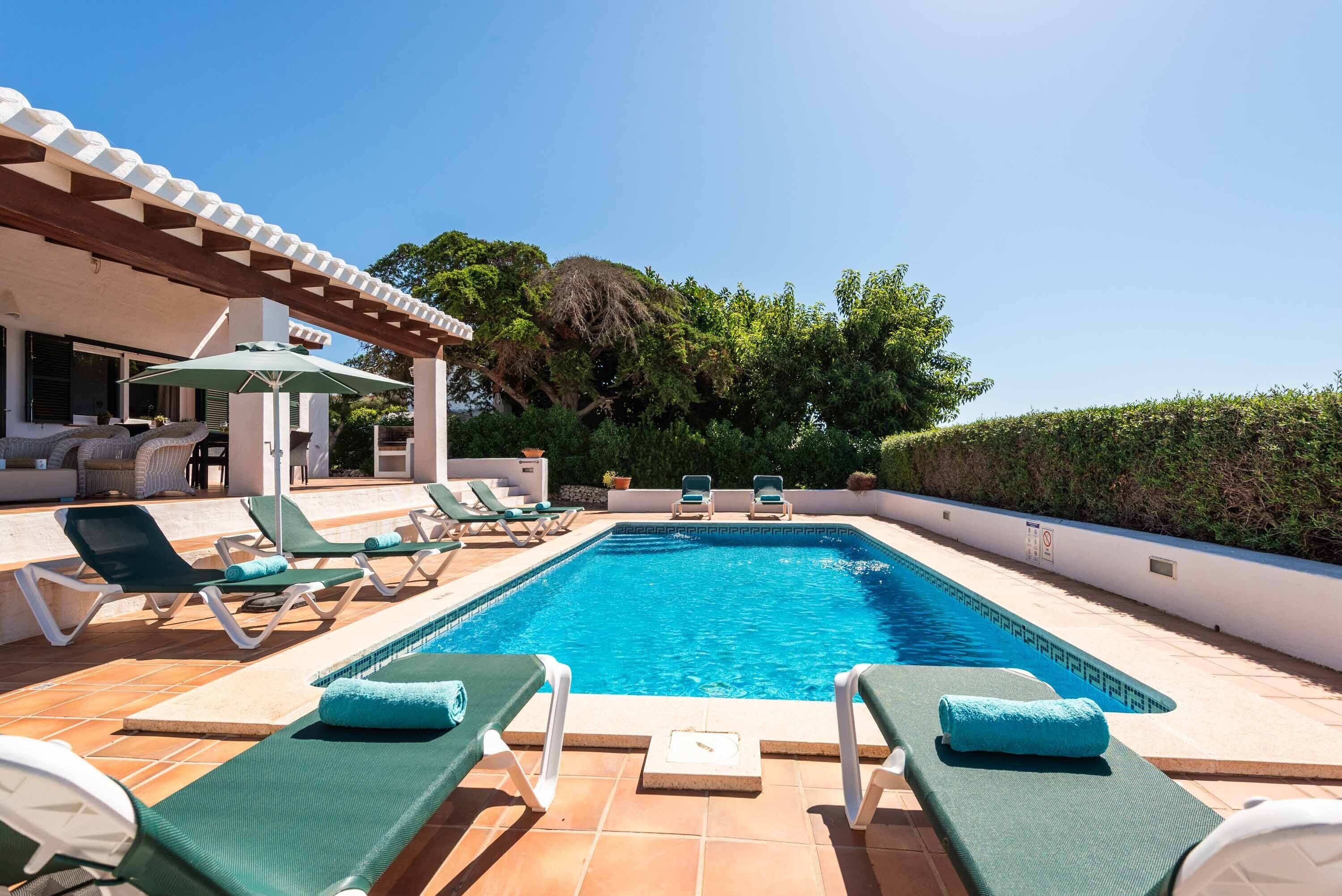 Piscina con sillas de descanso y sombrillas, rodeada de un jardín ajardinado y un edificio con pérgola.
