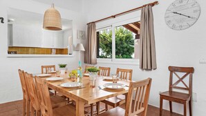 Een eetgedeelte met een houten tafel voor vier personen, omringd door houten stoelen. Boven de tafel hangt een lamp en rechts is een wandklok zichtbaar.