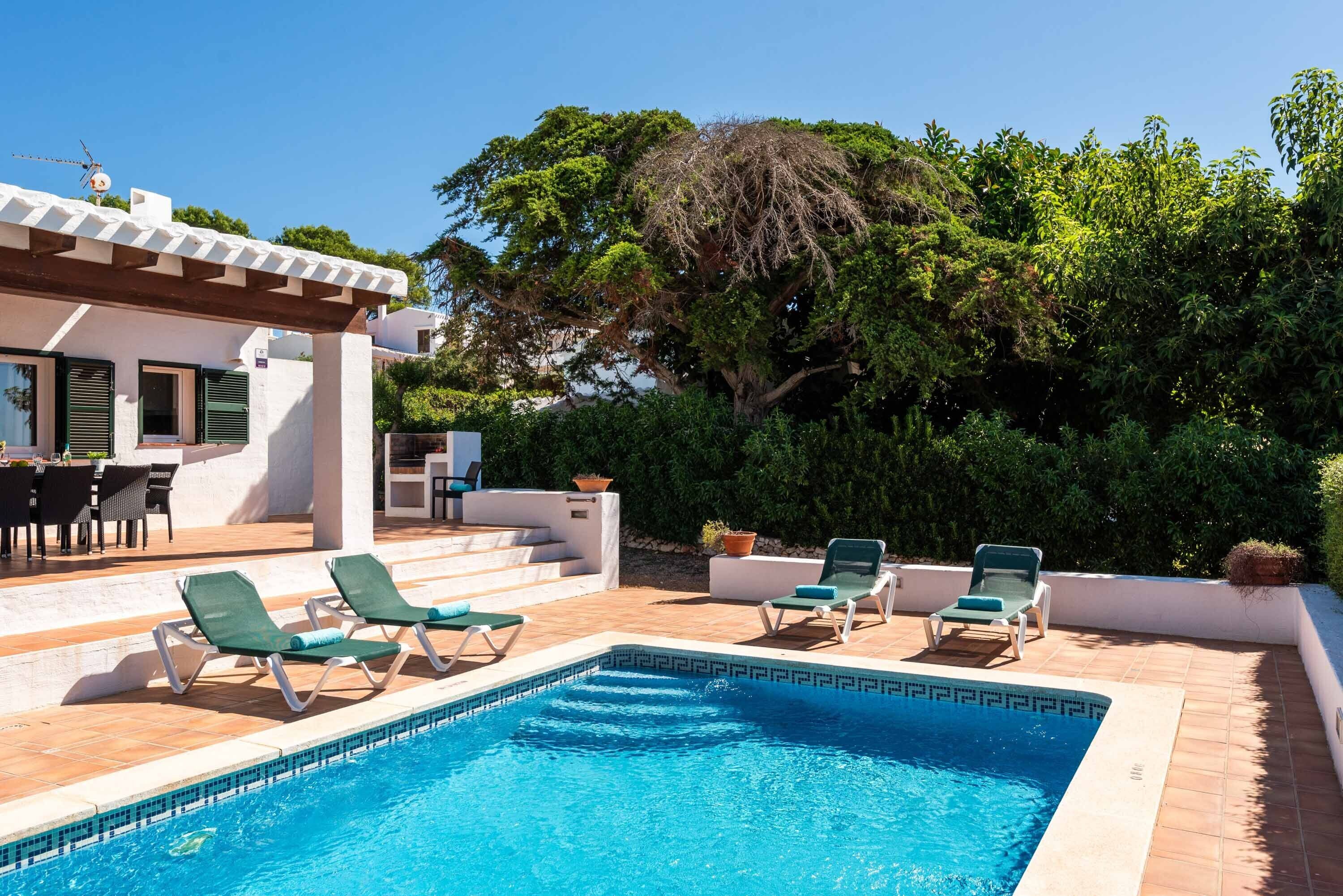 Un espace piscine aménagé avec des chaises longues, une pergola et une vue sur une maison à la façade blanche et aux volets verts.