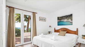 Een slaapkamer met een bed, een nachtkastje, een lamp, een afbeelding van een kustlandschap en uitzicht op palmbomen en de zee door een schuifdeur.