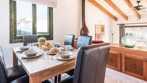 A dining area with a wooden table set for four, surrounded by chairs, with a view of the outdoors through large windows. - Es Llaut - Three Bedroom Villa, Sleeps 6 (Binibeca)