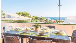 A terrace with wicker furniture, a table set for a meal, and a view of the sea and buildings. - Es Llaut - Three Bedroom Villa, Sleeps 6 (Binibeca)