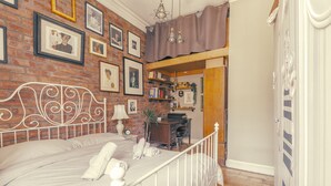 A bedroom with a brick wall, a white metal bed frame, a desk, and shelves with books and decorative items.