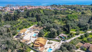 Luchtbeeld van een kustplaats met een opvallende villa, een zwembad, weelderige begroeiing en op de achtergrond bergen en een groot wateroppervlak.