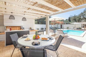 Een terras buiten met een ronde tafel, stoelen, een zwembad en een overdekt gebied met een barbecue.