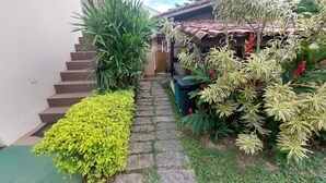 A stone pathway leading to a house with a tiled roof, surrounded by various plants and a green trash bin. - Plus Couple Suite with mini kitchen (Ilhabela)
