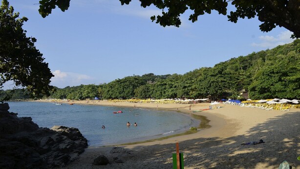 Ein Strand mit klarem Wasser, sandigem Ufer und einer bewaldeten Hügelkette im Hintergrund.